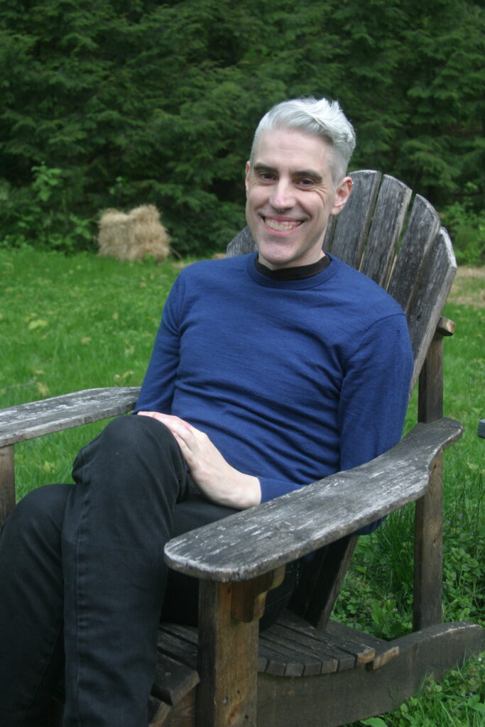 A picture of Rev. Ben Bigney, sitting in a weathered wooden chair with trees in the background.